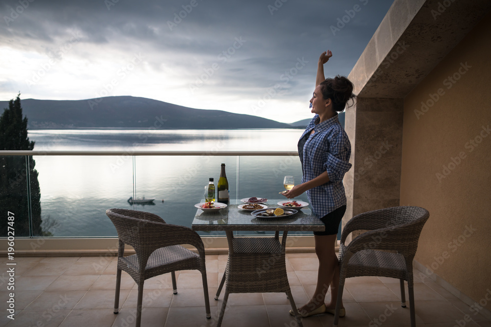 custom made wallpaper toronto digitalYoung happy woman is standing on the balcony of apartment near the table with food and drinks and waving to somebody. Smiling girl with glass of wine is looking on the beautiful sea and mountains
