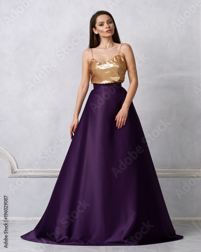 Charming brunette woman dressed in formal maxi dress with top decorated with golden sequins and purple satin bottom. Female model posing in elegant evening gown against white wall on background.