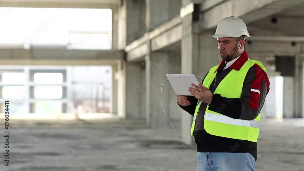Builder talks via tablet pc on project site. Worker with tablet computer on construction site. Workman in white hard hat holds in hand personal computer, looks in display Builder talks via tablet pc on project site. Worker with tablet computer on construction site. Workman in white hard hat holds in hand personal computer, looks in display
