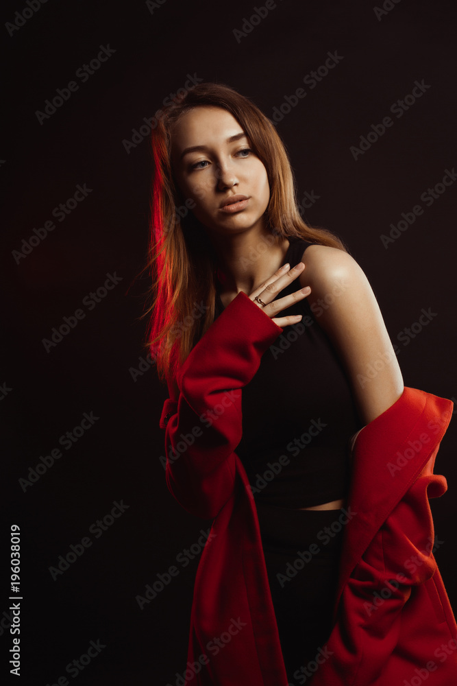 Sensual young woman with natural makeup and shiny hair posing in trendy red coat at studio