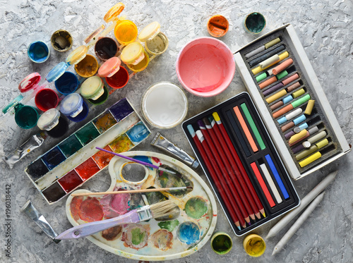 Artistic tools for drawing paintings on a gray concrete background. Palette, gouache, oil paint, brushes, colored crayons, pastel, colored pencils. Top view..