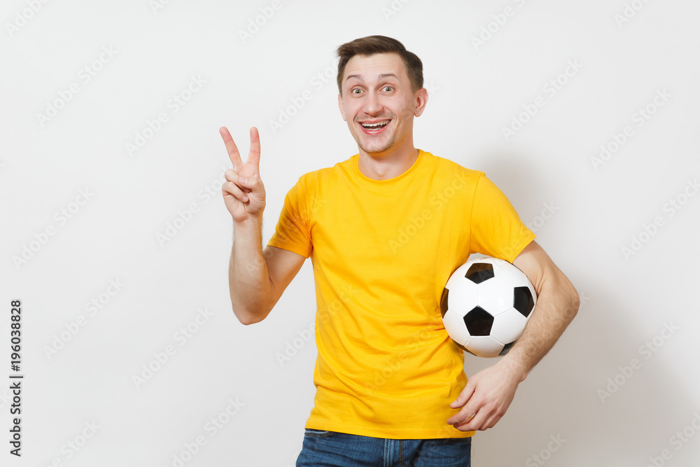 Young cheerful European man, fan or player in yellow uniform hold soccer ball, show victory sign, cheer favorite football team isolated on white background. Sport, play football, lifestyle concept. Young cheerful European man, fan or player in yellow uniform hold soccer ball, show victory sign, cheer favorite football team isolated on white background. Sport, play football, lifestyle concept.