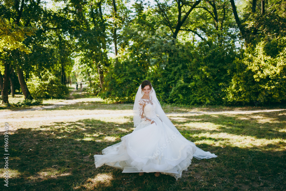 Beautiful wedding photosession. The smiling bride in a white lace dress with a plume, long sleeves, hairpin in hair, white veil turns around in a large green garden on weathery sunny day Beautiful wedding photosession. The smiling bride in a white lace dress with a plume, long sleeves, hairpin in hair, white veil turns around in a large green garden on weathery sunny day