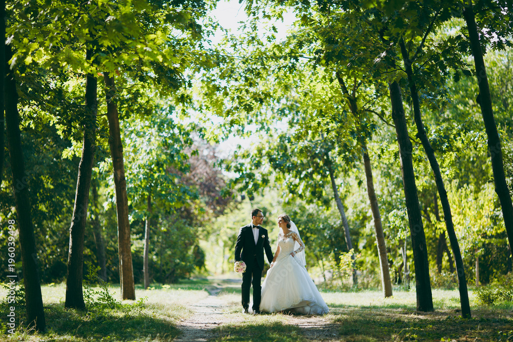 Fototapeta premium Beautiful wedding photosession. The groom in a black suit with bouquet and his bride in a white lace dress with a long plume and veil smile and walk along the path in large green garden on sunny day