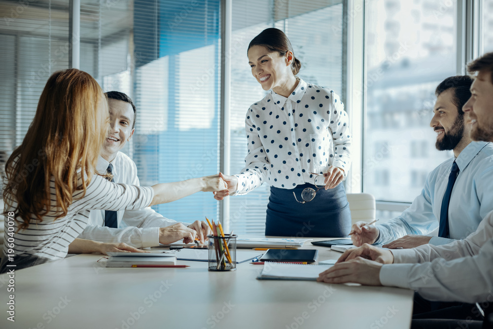 Pleasant cooperation. Upbeat young female boss shaking hands with her female colleague, congratulating her with promotion, while carrying out a meeting - obrazy, fototapety, plakaty 