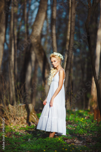 blond woman stands in white dress by thin tree.