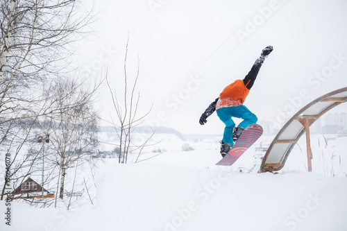 Photo from back of young athlete skating on snowboard with springboard against background of trees