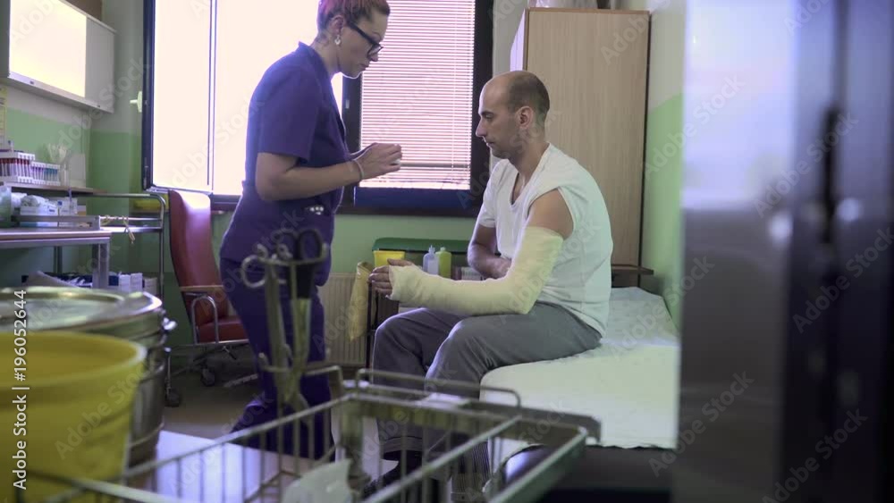 Nurse in uniform bandaging injured hands of male patient, he sits on bed in hospital room and she putting sticking plaster on bandage at his arm, sterile tools on medical table close up, dolly shot