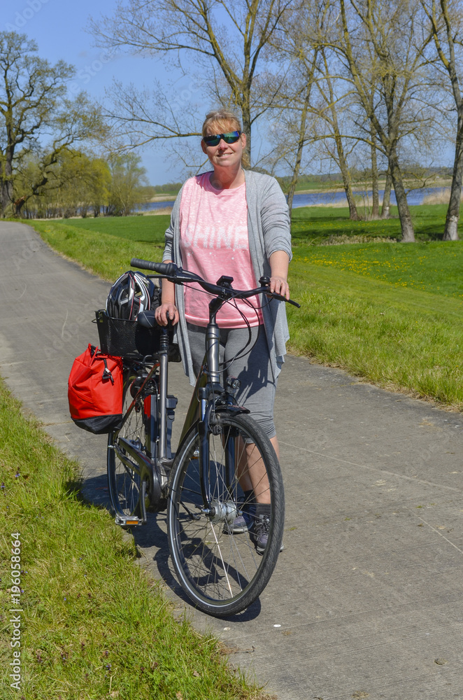 Obraz premium Woman standing on a bikeway beside her bike in rural surrounding near the Elbe River in the Lueneburg Heath at a sunny day with blue sky - portrait photo
