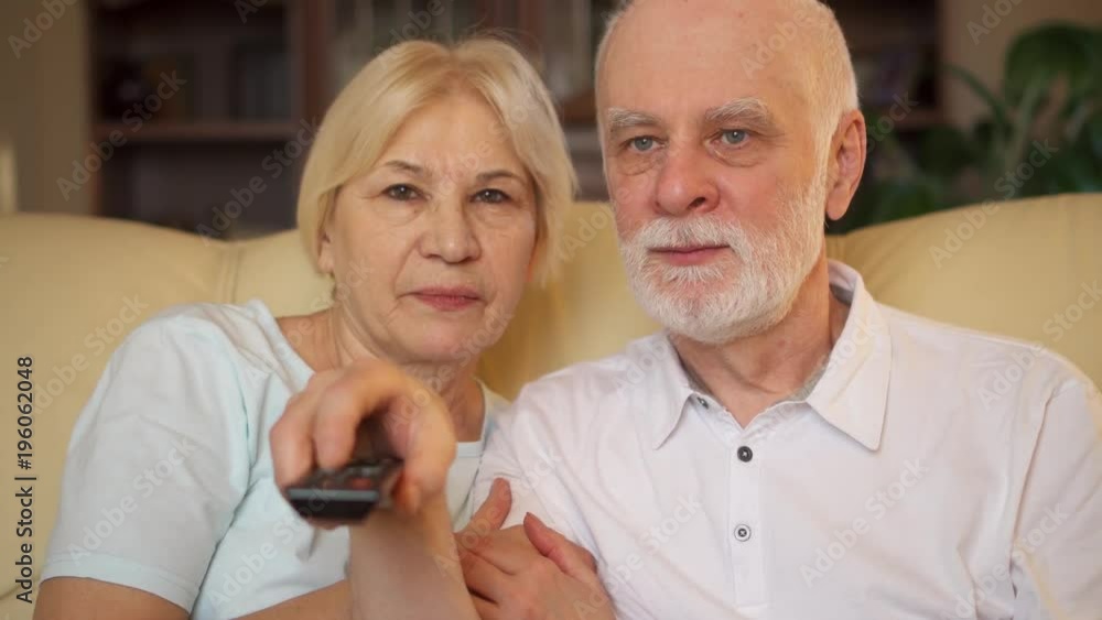 Senior couple relaxing on sofa at home. Happy family enjoying time together watching TV. Pensioners switching channels with remote control Senior couple relaxing on sofa at home. Happy family enjoying time together watching TV. Pensioners switching channels with remote control