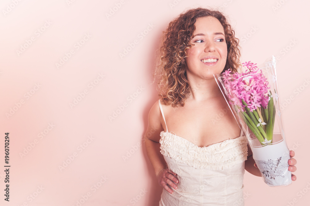 Beautiful curly woman in white dress with hyacinth flowers in hands on a light pink background. Hello spring concept. Beautiful curly woman in white dress with hyacinth flowers in hands on a light pink background. Hello spring concept.
