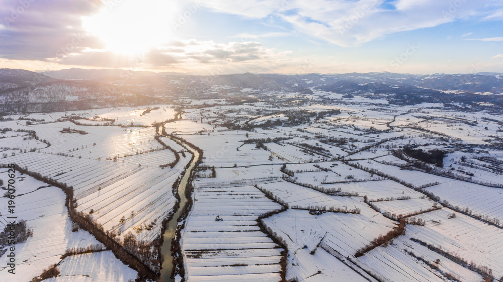 Obraz premium Aerial view of river flowing through snow covered countryside at sunset.