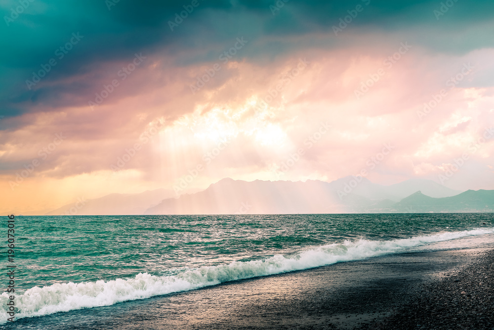 Beautiful summer seascape with mountains silhouette. Cloudy pink sky with sunrays through clouds. Salerno beach, Italy
