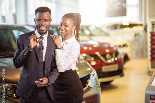 Visiting car dealership. Beautiful african couple is holding a key of their new car, looking at camera and smiling photo