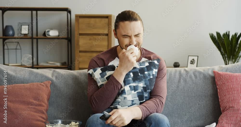 Portrait of Caucasian young man crying while sitting on the sofa and hugging a pillow in the cozy room. watching a melodrama. Indoor Portrait of Caucasian young man crying while sitting on the sofa and hugging a pillow in the cozy room. watching a melodrama. Indoor