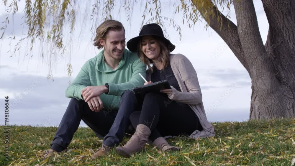 Young couple sitting under tree looking at tablet and talking and smiling with each other. Young couple sitting under tree looking at tablet and talking and smiling with each other.