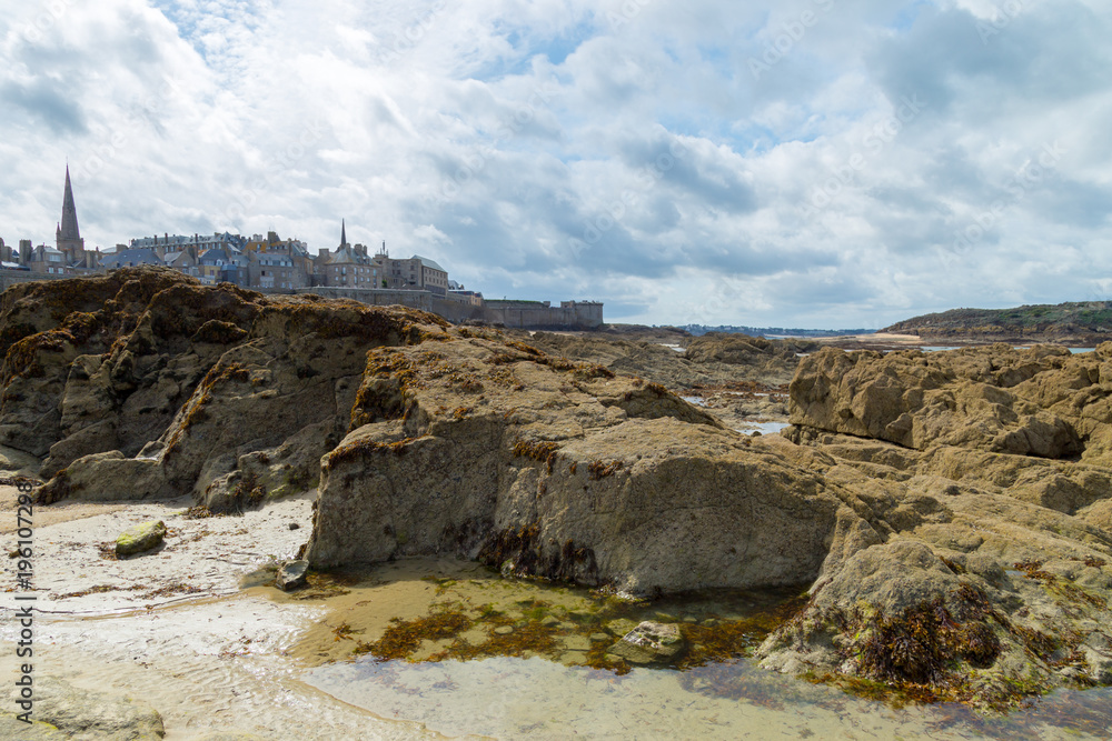 Fototapeta premium Sea rocks and beach shore with Saint Malo city Brittany, Europe on the background