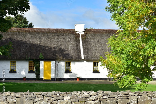 White country cottage with high thatched roof and bright yellow door and window frames behind low stone wall.