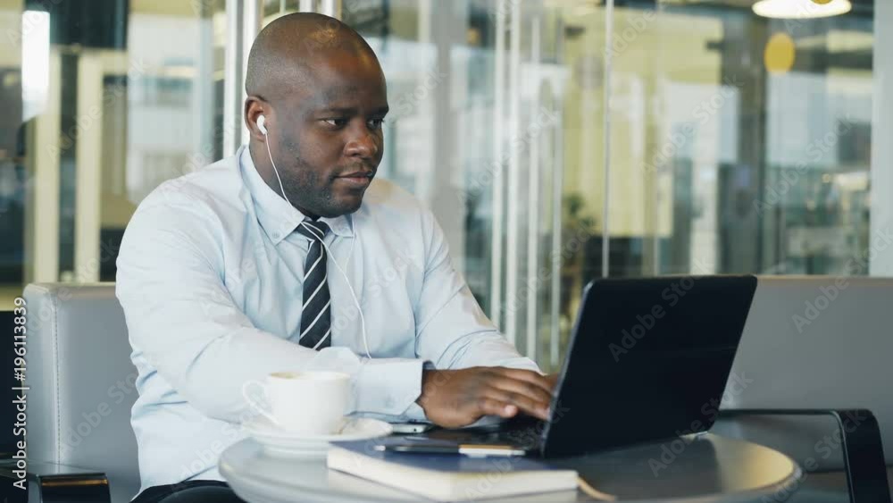 African American businessman in formal wear working and surfing social media on laptop while listening to music with earbuds in his ears in a glassy cafe African American businessman in formal wear working and surfing social media on laptop while listening to music with earbuds in his ears in a glassy cafe