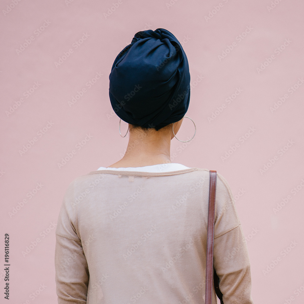 Image of a Muslim Malay woman standing and facing on a plain pink background. She is wearing a blue turban and a classy very outfit.