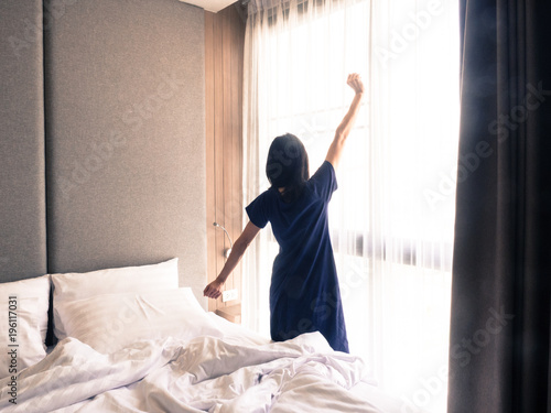 Woman stretching in bed after waking up, back view. Woman sitting near the big white window while stretching on bed after waking up with sunrise at morning, back view.