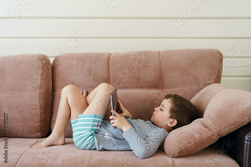 cute boy in casual clothes lying on the couch watching and playing tablet in the house