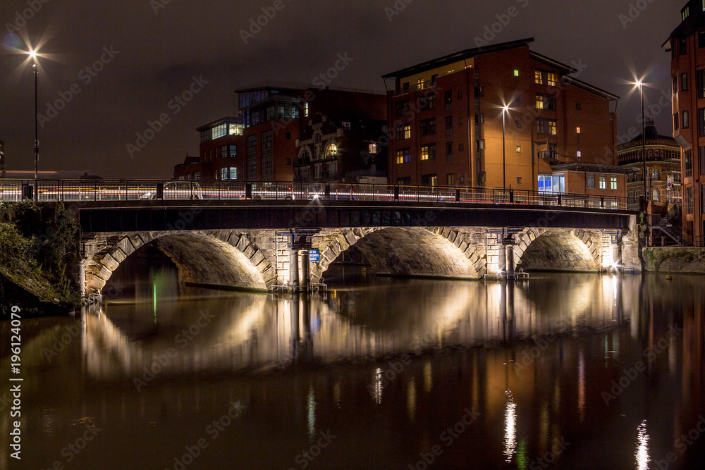 Night scene with lit bridge over a still river with long reflections in a residential area with low rise modern buildings.