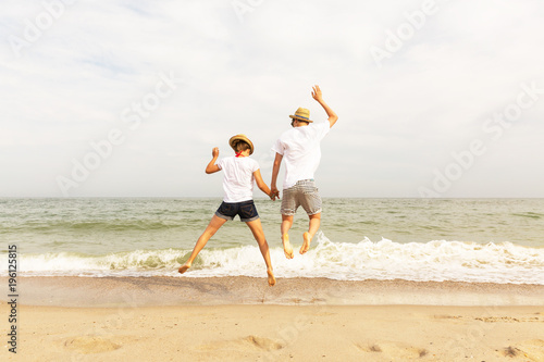 Two teenagers: a girl and a boy with blond hair, dressed in white T-shirts and shorts jumping up holding hands on a sea beach. Copy space.