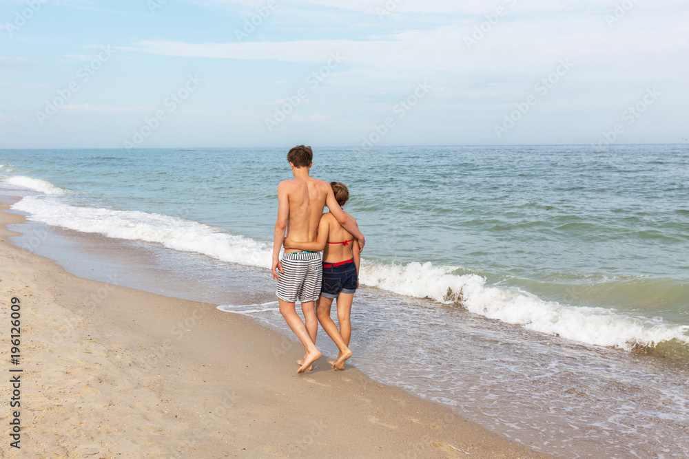 Two teenagers: a girl and a boy with blond hair, dressed in a swimsuit are walking on a sea beach. Back view. Two teenagers: a girl and a boy with blond hair, dressed in a swimsuit are walking on a sea beach. Back view.