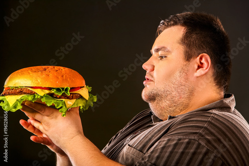 Man eating fast food hamberger. Fat person made great huge hamburger and admires him, intending to eat it. Junk meal leads to obesity. Health problems due to malnutrition.