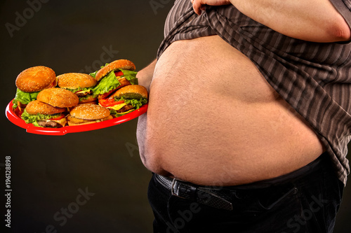 Man belly fat with hamberger group fast food. Breakfast for overweight person. Cropped shot of junk meal leads to obesity. Male lifted shirt, shows bare stomach. Health problems due to malnutrition.
