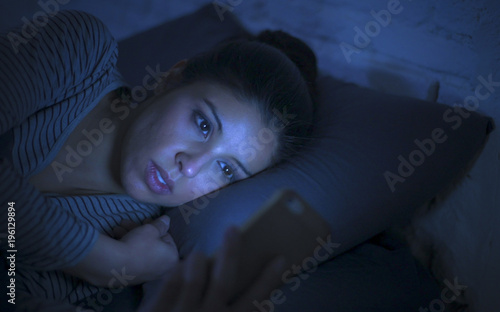 portrait of young beautiful latin woman using mobile phone late night sleepless lying in bed in the dark in smartphone and internet dating app addiction