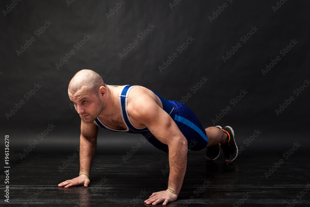 A young athletic man in blue wrestling tricky and blue shorts does push-ups on a black-haired isolated background in a photo studio A young athletic man in blue wrestling tricky and blue shorts does push-ups on a black-haired isolated background in a photo studio