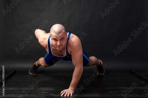A young athletic man in blue wrestling tricky and blue shorts does push-ups on a black-haired isolated background in a photo studio