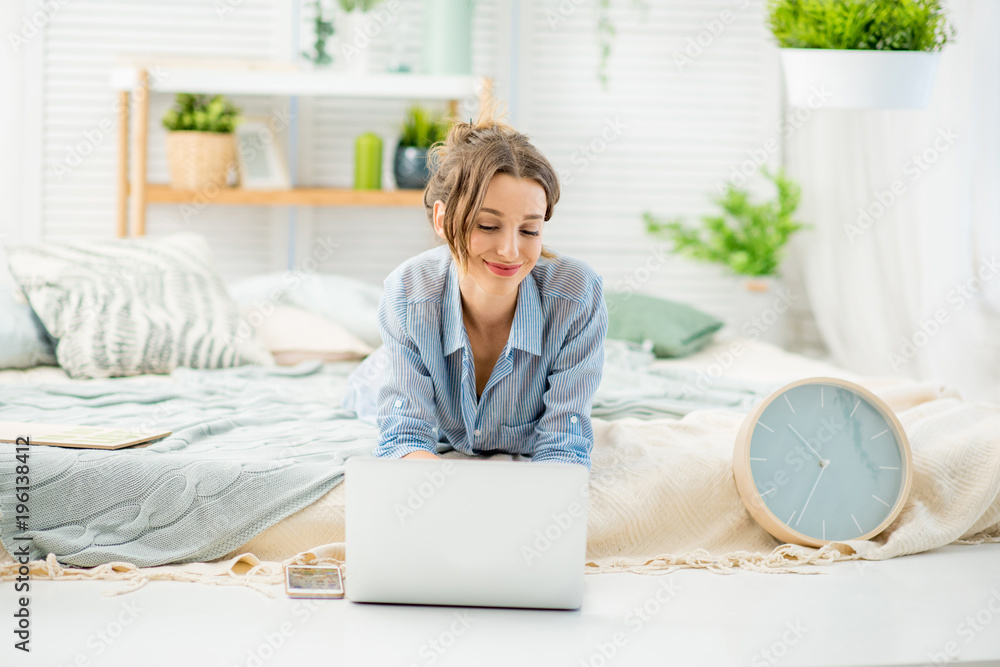 Young woman in blue shirt working with laptop lying on the bed in the bright and cozy bedroom at home Young woman in blue shirt working with laptop lying on the bed in the bright and cozy bedroom at home
