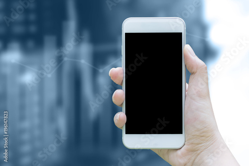 Mockup image of hand holding white mobile phone with blank black display on graph and stock background blurred. Concept is technology of business and financial.