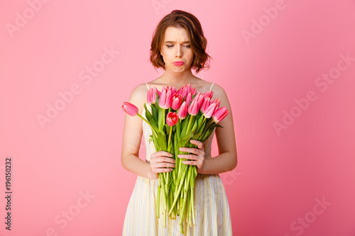 portrait of upset young woman with bouquet of pink spring flowers isolated on pink