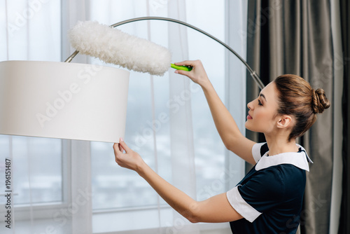 young beautiful maid in uniform cleaning torchere with duster photo