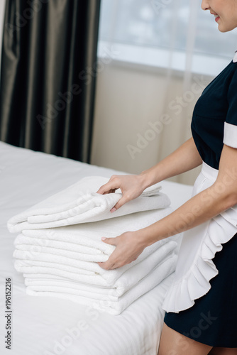 cropped shot of smiling maid in uniform with stack of clean towels on bed in hotel suite