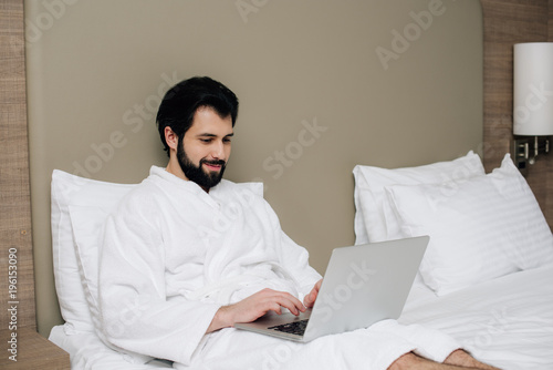 handsome man in bathrobe using laptop in bed at hotel suite