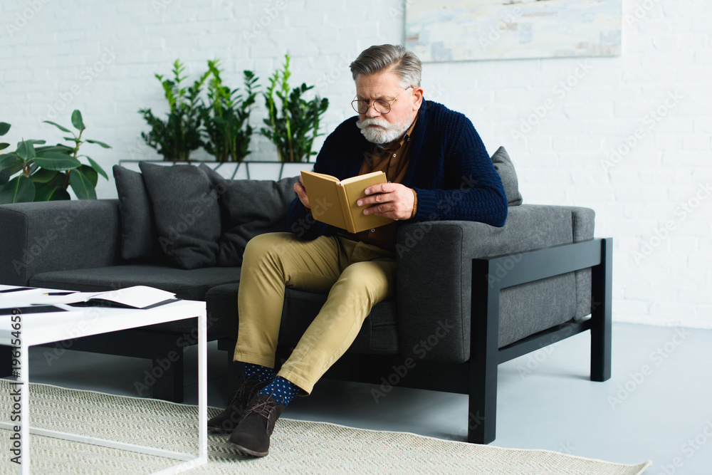 serious bearded senior man sitting on sofa and reading book at home