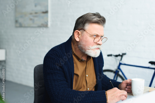 thoughtful senior man in eyeglasses holding cup and taking notes at home