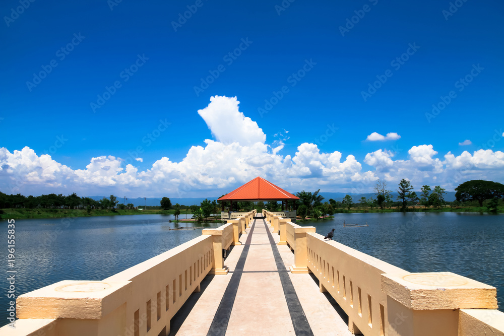 The orange triangular pavilion is located in the middle of the water, with a bridge in the pathway and white clouds, with a beautiful blue sky as the background.