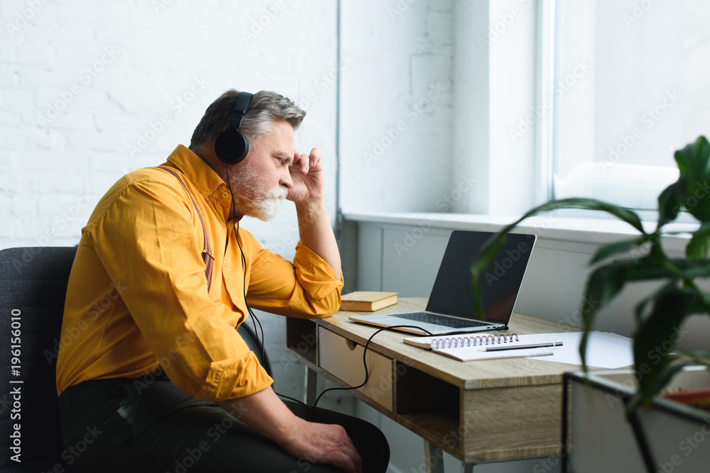 side view of serious senior man in headphones using laptop with blank screen at home side view of serious senior man in headphones using laptop with blank screen at home