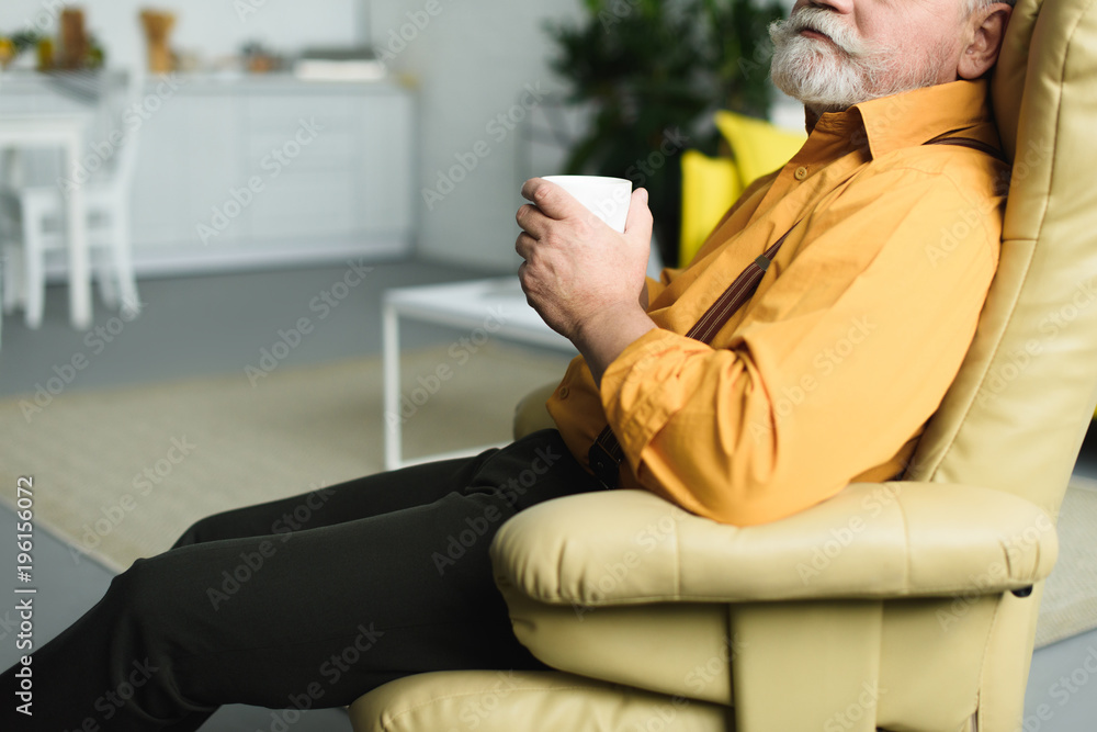 cropped shot of bearded senior man holding cup and sitting on armchair at home