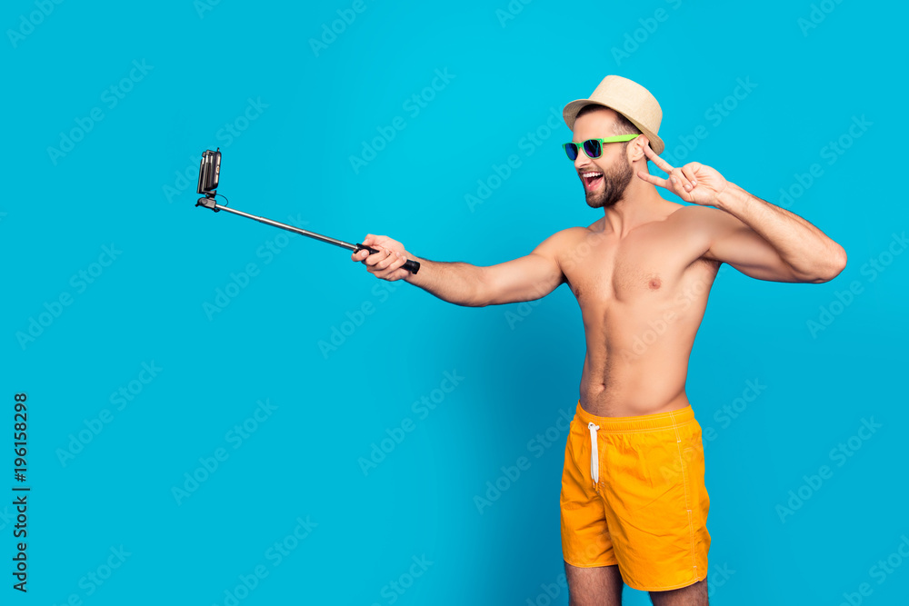 Laughing, attractive, stylish, muscular, ladies' man using selfish stick with smart phone, shooting self picture, having video call, gesturing v-sygn, isolated on blue background Laughing, attractive, stylish, muscular, ladies' man using selfish stick with smart phone, shooting self picture, having video call, gesturing v-sygn, isolated on blue background