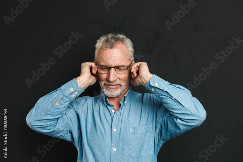 Image of intelligent gentleman 50s wearing businesslike outfit covering ears due to annoying noise or interlocutor, isolated over black background