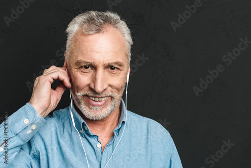 Portrait of handsome pleased mature man 60s with gray hair looking on camera while listening to music via white headphones, isolated over black background