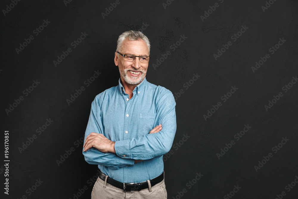 Portrait of caucasian male pensioner 60s with grey hair and beard smiling and looking aside while standing with arms folded, isolated over black background