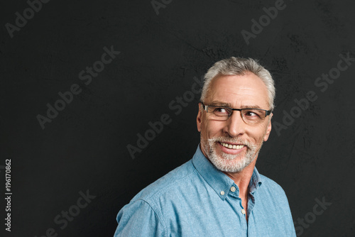 Horizontal portrait of mature unshaved man 60s with grey hair wearing eyeglasses smiling and looking aside on copyspace, isolated over black background